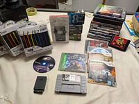 Lot shows assorted video games and accessories including DVC cassette tapes, Nintendo DS stylus pens, and selected game discs and cartridges spread out on a table.