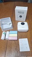 Photo showing the printer box, portable mini printer unit, USB cable, four rolls of thermal paper, instruction manual on wood surface