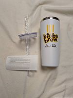 Side view of white Biebs Brew tumbler with silver rim, yellow stripes, and Tim Hortons x Justin Bieber logo. Clear plastic straw and spare lid wrapped in plastic beside tumbler on fabric background.