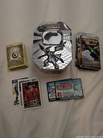 Photo showing the opened Pokemon Black & White 2011 metal tin, a stack of Pokemon cards, some empty card wrappers, and sports cards on a light background.