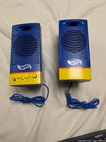 Front view of two Hot Wheels computer speakers, blue with yellow base, both feature the Hot Wheels logo and front speaker grills. The left speaker has control knobs and a power button.