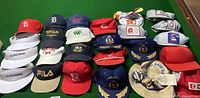 Multiple men's sports caps and hats including branded visors, team baseball caps, and novelty hat arranged on green surface.