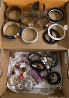 Overall lot of bracelets including metal cuffs, wooden and plastic bangles, and multiple beaded bracelets arranged in a cardboard box.