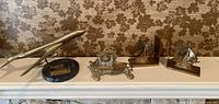 Brass airplane on black marble base with brass plaque, ornate brass inkwell with glass insert, and brass sailboat bookends on mantle against floral wallpaper.