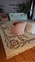 Large area rug with floral pattern border and fringe edges, displayed under white and pink upholstered furniture, hardwood floor visible
