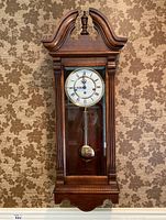 Full view of Hentschel wall clock hanging against floral wallpaper showing wooden case, glass front, dial and pendulum.