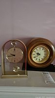 Both clocks shown side by side on a mantel surface, one in wood frame and the other in clear acrylic frame.