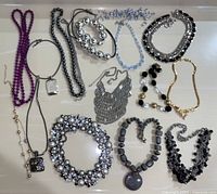 Photo showing assortment of 11 costume necklaces of various styles, colors and materials (stone, metal, plastic) laid out on a white surface.