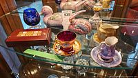Photo showing multiple items on glass shelf including lusterware teacups, decorative birds, and a red box.