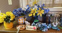 Variety of faux flowers in yellow and blue tones, ceramic vases including one with sunflower design, plaques labeled Grandpa and Grandma, and blue glass candlesticks