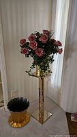 Full view of artificial pink roses set on a shiny gold-tone mirrored plant stand with a square base and adjustable gold sections, alongside a separate gold plastic pot on the floor.