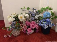 Collection of faux flowers in different vases and pots arranged on red carpet against white wall
