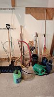 Gardening tools and accessories arranged indoors including shovel, saws, rakes, watering cans, plant sprayers, boots