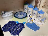 Overview photo showing all kitchenware items together on a countertop, highlighting the goose-themed ceramic canisters, dishes, figurines and blue trivets and cutting boards.