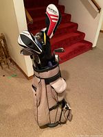 Golf bag with clubs inside shown standing upright indoors on carpet near stairs.