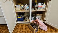White cabinet with open doors showing shelves filled with multiple hardware and home maintenance product containers. Boxes and bags of items placed on the floor in front.