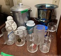 Photo showing hammered aluminum ice bucket, Buddha figure, silver plated urn and various glassware including beer tankards and cups