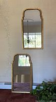 Two gold toned framed wall mirrors, one mounted on the wall and one propped against it. Both show rectangular reflective surfaces with decorative frames.