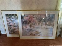 Two framed landscape art prints leaning against wall on carpet, showing soft pastel scenes with water, rocks, and flowering trees.
