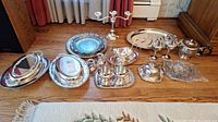 Full view of silverplated serving ware including trays, goblets, small dishes, candelabra, and candlesticks arranged on wood floor.