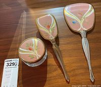 Photo of entire set showing hand mirror, hairbrush, and powder jar with matching decorative lids on wooden surface.