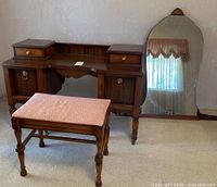 Front view of vintage wooden vanity with two small drawers on top, two side cabinets, and matching cushioned stool in front. Large mirror is leaning against wall.