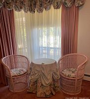 Overview of two wicker chairs with cushions flanking a round wicker table draped in cloth in front of window