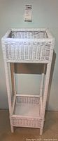 Front view of the white wicker two-tier plant stand showing the square top shelf with raised edges and long narrow legs covered in wicker.