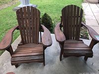 Front angle view of both brown wooden Adirondack chairs on a concrete patio with grass in background