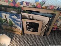 Multiple vintage paintings and photographs stacked together on bed, including signed paintings and black-and-white photographs with cardboard mounts.