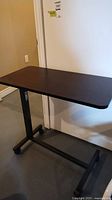 Side view of rolling adjustable bed table showing dark wood tabletop and black metal frame with wheels on base.