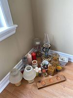 Wide angle photo showing all candle lamps, votives, tea lights, hurricane lamp, and decorative candle balls arranged in a corner on a wood floor near a window and wall.