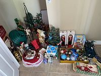 Wide view showing collection of small decorated faux Christmas trees, nutcrackers, holiday figurines, assorted ornaments, and storage boxes gathered on a tiled floor.