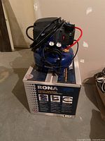 Rona RCK2-4 Air Compressor sitting on its original box, showing full unit and hose wrapped around top.