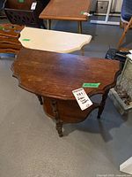 Wooden demilune table with dark brown finish, scalloped top edge, carved legs, and lower shelf, accompanied by a painted white demilune table in the background.
