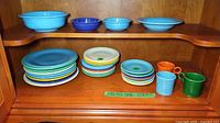 Overall view of two shelves showing stacked plates, bowls and cups in assorted colors.