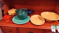 Shelf view showing covered serving dish (yellow/ivory), teal pitcher, cobalt blue small pitcher/creamer, orange handled bowl, teal plate, and two yellow/ivory plates