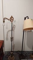 Full view of two vintage floor lamps side by side, one with beige lampshade, the other with decorative scroll arm and bare bulb.