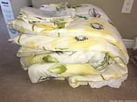 Stack of king size bed linens and bedspread folded together on carpet, showing floral pattern in yellow, green and gray on white background.