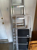 View showing both the 6-foot aluminum step ladder and the 3-foot metal framed step ladder leaning in a garage near a door, giving perspective on size and materials.