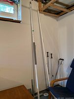 Two tall white adjustable Healthcraft Superpoles leaning against a basement wall near a window, with gray grip sections and weighted black bases.