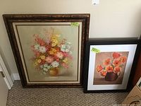 Two framed floral artworks side by side on floor - larger floral oil painting in 30x26 frame with bouquet of daisies and other flowers, signed in lower right. Smaller floral poppy print in simple black frame.
