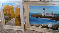 Two framed paintings: one autumn road scene with yellow trees, one coastal lighthouse scene with seashells.