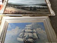 Two framed paintings stacked: top painting is a landscape, bottom is a ship at sea.