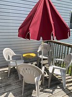 Wide angle view showing all four white plastic chairs arranged around a yellow metal side table with a red patio umbrella overhead on a wooden deck.