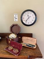 Overview of vintage eyeglasses, Parker pens, Bulova Cigna clock in wooden case, and black-framed battery wall clock on wooden surface.