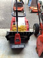 Front view of red and black Toro Power Shovel with yellow extension cord wrapped on handle. Other lawn equipment visible behind.