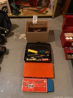 Overall view of the lot including a cardboard box with tools, a black hand tools case, and an orange metal ratchet socket set case on a concrete floor.