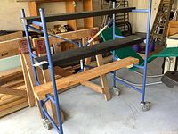 Full view of the blue metal scaffolding frame with two black platforms and wooden plank, wheels visible on legs.