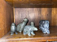 Five carved soapstone animal figurines displayed on a wooden shelf. Figures include a bird, seal, polar bear, and two crouching animals that resemble cats or bears. The stones are polished and predominantly dark gray.